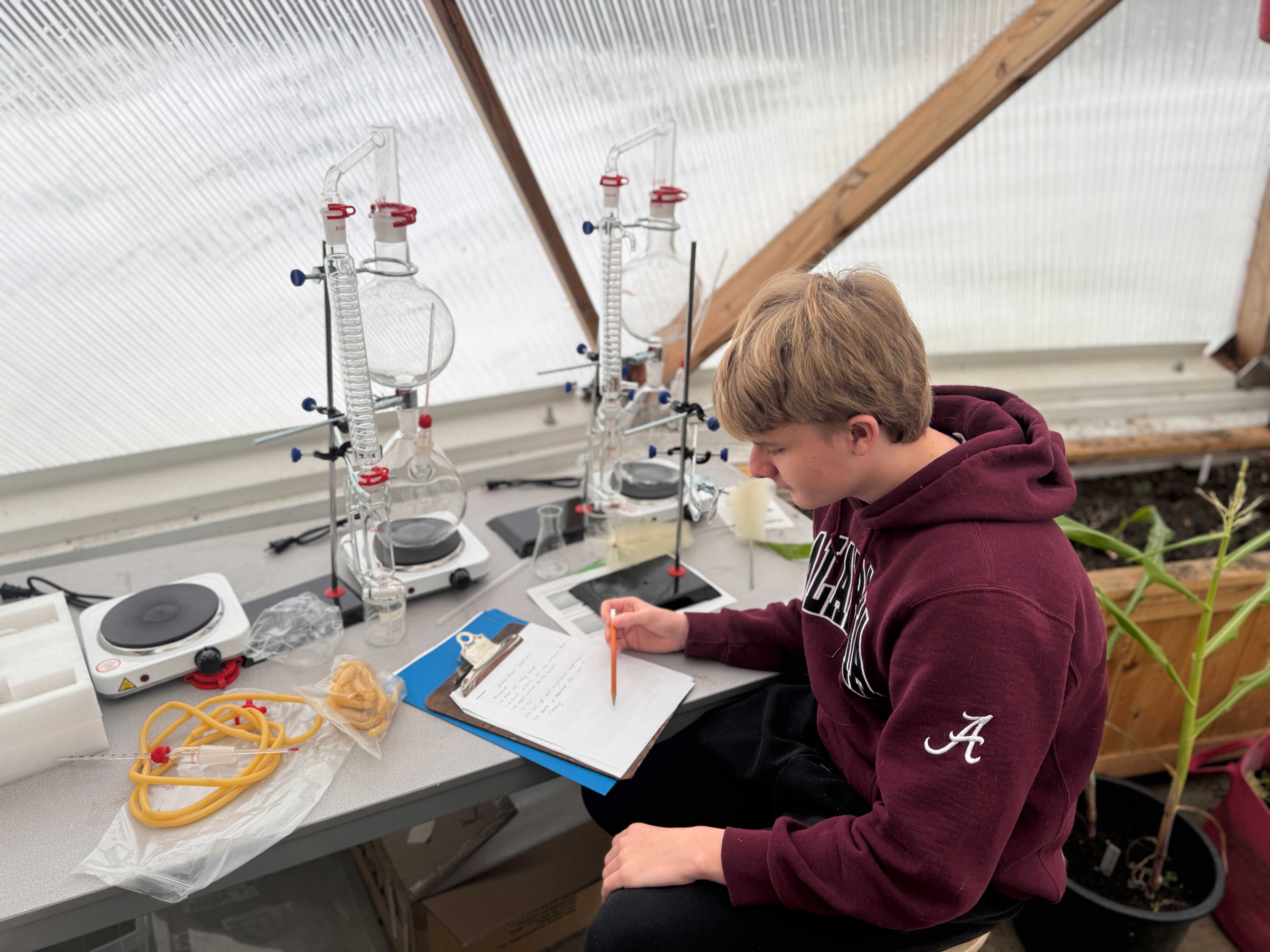 Student testing samples in the SFDCI dome, working at a table equipped with glass distillation apparatus, hot plates, and notes on a clipboard, surrounded by a greenhouse-like structure with natural light.